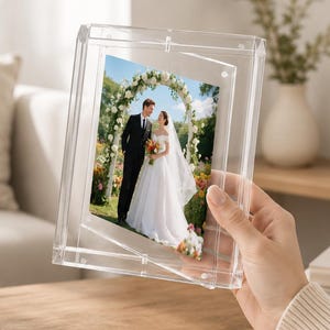 Peut inclure: Un cadre photo flottant en acrylique transparent au design rectangulaire, contenant une photographie d'un couple en tenue de mariage sous une arche fleurie. Le cadre présente une construction à double paroi créant un effet de suspension.
