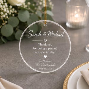 Peut inclure: Un ornement circulaire en acrylique transparent avec un bord facetté, suspendu par un cordon doré. Le texte indique : Sarah & Michael, Thank you for being a part of our special day!, With love, The Newlyweds, May 18, 2025, avec deux petits cœurs.