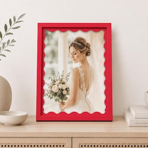 Peut inclure: Un cadre photo rectangulaire avec une finition rouge vif et un bord intérieur festonné. Le cadre contient une photographie d'une personne portant une robe de mariée en dentelle blanche, tenant un bouquet de fleurs blanches et un voile.