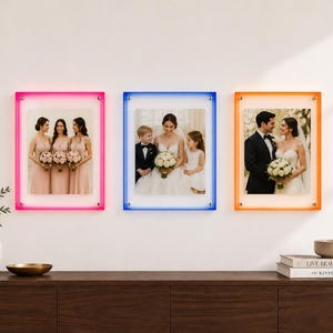 May include: Three rectangular wall-mounted photo frames with neon-style borders in hot pink, bright blue, and vibrant orange. Each frame features a clear acrylic panel held by silver hardware, displaying wedding-themed photographs of people.