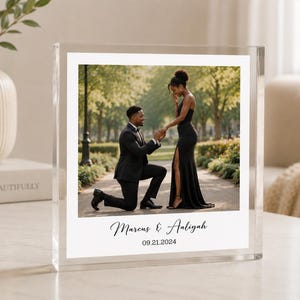 Peut inclure: Un cadre photo en bloc acrylique transparent présentant une photographie imprimée d'une personne en costume noir agenouillée pour demander en mariage une personne en robe noire. Sous la photo, le texte indique Marcus & Aaliyah et la date 09.21.2024.