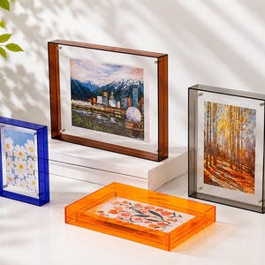 Pode incluir: Quatro molduras retangulares de acrílico nas cores azul, castanho, laranja e cinzento. A moldura azul contém uma impressão de margaridas, a castanha mostra um horizonte urbano, a laranja uma ilustração floral e a cinzenta uma floresta.