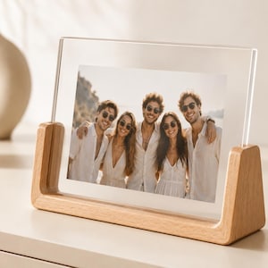Peut inclure: Un cadre photo en acrylique transparent soutenu par une base en bois clair. Le cadre contient une photographie de cinq personnes debout ensemble à l'extérieur, portant des vêtements blancs et des lunettes de soleil, posé sur une surface plane.