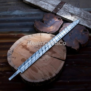May include: A silver-coloured, patterned sword blade with a pointed tip. The blade has a textured design and is displayed on a wooden surface. The blade is angled across a wooden log and other wooden pieces.
