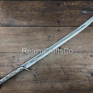 May include: A silver-colored sword with a curved blade and ornate handle. The blade features etched designs, and the handle has a detailed pattern. The sword is displayed on a wooden surface, with the text "RealmCraftsCo" visible.