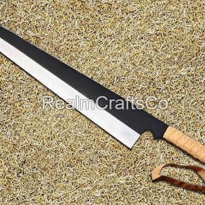 Op de afbeelding: Een groot mes met een zwart en zilveren lemmet, een bruin omwikkelde handgreep en een leren riem. Het lemmet is ongeveer 61 cm lang. De tekst "RealmCraftsCo" is zichtbaar op het lemmet.