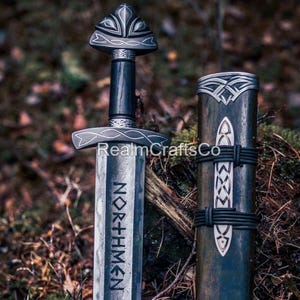 May include: A Viking sword and sheath are displayed. The sword has a silver crossguard and pommel, a dark wooden handle, and the word "NORTHMEN" etched on the blade. The sheath is dark with silver detailing.