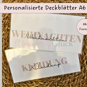 Könnte beinhalten: Zwei rechteckige A6-Einlegeblätter aus weißem Papier mit glänzender Oberfläche. Das obere Blatt zeigt den Text "Weihnachten" in großen hellbraunen Buchstaben mit "Umschlag Glück" in Schreibschrift. Das untere Blatt zeigt das Wort "Kleidung".