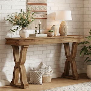May include: A wooden console table with a natural finish, featuring an X-shaped leg design. The table is adorned with a vase of flowers, a lamp, candles, and decorative items. A potted plant and woven basket add to the decor. The table is 60 inches long.