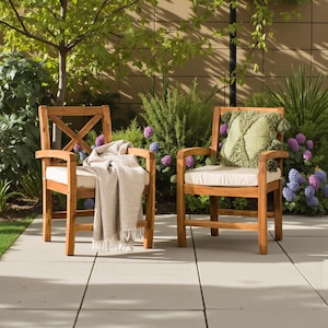 May include: Two wooden outdoor armchairs with cream-colored cushions sit on a patio. One chair has a beige throw blanket draped over it, and the other has a green decorative pillow. The chairs are surrounded by lush greenery and flowers.