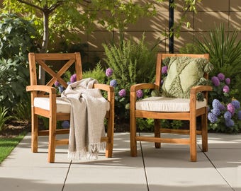 May include: Two wooden outdoor armchairs with cream-colored cushions sit on a patio. One chair has a beige throw blanket draped over it, and the other has a green decorative pillow. The chairs are surrounded by lush greenery and flowers.