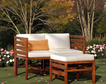 May include: Outdoor wooden sectional sofa with white cushions and an orange lumbar pillow. A matching wooden ottoman with a white cushion sits in front of the sofa. The furniture is on a green surface with a backdrop of trees and flowers.