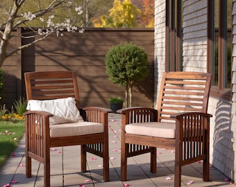May include: Two wooden outdoor armchairs with beige cushions are on a patio. The chairs have a slatted back and armrests. A white pillow with a geometric pattern rests on one chair. The chairs are surrounded by greenery and a wooden fence.