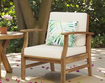 May include: Wooden outdoor armchair with cream-colored cushions and armrests. A decorative pillow with a green and white leaf pattern rests on the chair. A small round table and potted plant are visible in the background.