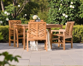 May include: A wooden outdoor dining set with a table and four chairs. The chairs have a chevron design on the backrests. A white blanket is draped over one chair. The set is on a stone patio, surrounded by greenery.