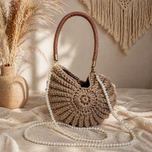 May include: A handcrafted, brown crochet handbag with a braided brown handle and gold-colored hardware. The bag features a pearl beaded strap and a unique seashell design. The bag is displayed on a neutral-toned surface.