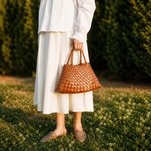K&ouml;nnte beinhalten: Eine braune, gewebte Ledertasche mit zwei Griffen. Die Tasche hat ein Korbgeflechtmuster und wird von einer Person gehalten, die ein wei&szlig;es Kleid und braune Schuhe tr&auml;gt. Der Hintergrund ist eine gr&uuml;ne Wiese mit kleinen wei&szlig;en Blumen.