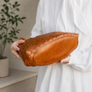K&ouml;nnte beinhalten: Eine handgefertigte Handtasche aus braunem Leder mit einem geflochtenen Muster. Die Tasche hat einen abgerundeten Boden und einen geflochtenen Henkel. Die Tasche wird von einer Person gehalten, die ein wei&szlig;es Hemd tr&auml;gt.