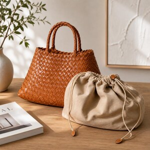 K&ouml;nnte beinhalten: Eine geflochtene braune Ledertasche mit geflochtenen Griffen steht neben einem beigefarbenen Kordelzugbeutel. Die Tragetasche hat eine Trapezform. Der Kordelzugbeutel ist mit einem Kordelzug und Lederakzenten verschlossen.