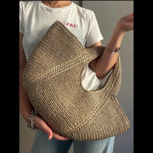 Peut inclure: Un sac fourre-tout tissé beige clair. Le sac a une grande poignée ouverte et une forme structurée. La texture du sac est visible, avec un motif de tissage détaillé. Le sac est tenu par une personne.