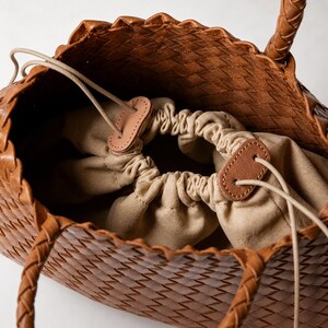 K&ouml;nnte beinhalten: Braune Leder-Tragetasche mit geflochtenem Design und geflochtenen Henkeln. Der Innenraum hat einen beigen Kordelzugverschluss und ein beiges Stofffutter. Die Tasche hat ein Korbgeflechtmuster.