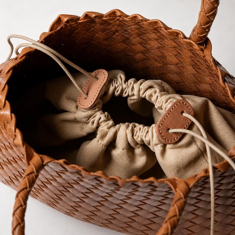 K&ouml;nnte beinhalten: Braune Leder-Tragetasche mit geflochtenem Design und geflochtenen Henkeln. Der Innenraum hat einen beigen Kordelzugverschluss und ein beiges Stofffutter. Die Tasche hat ein Korbgeflechtmuster.