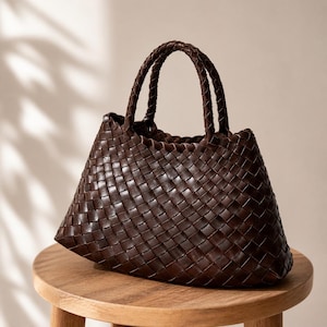 Peut inclure: Un sac fourre-tout en cuir tressé marron foncé avec des anses tressées. Le sac est de forme trapézoïdale et repose sur un petit tabouret en bois. Le motif de tissage est visible sur tout l'extérieur du sac.