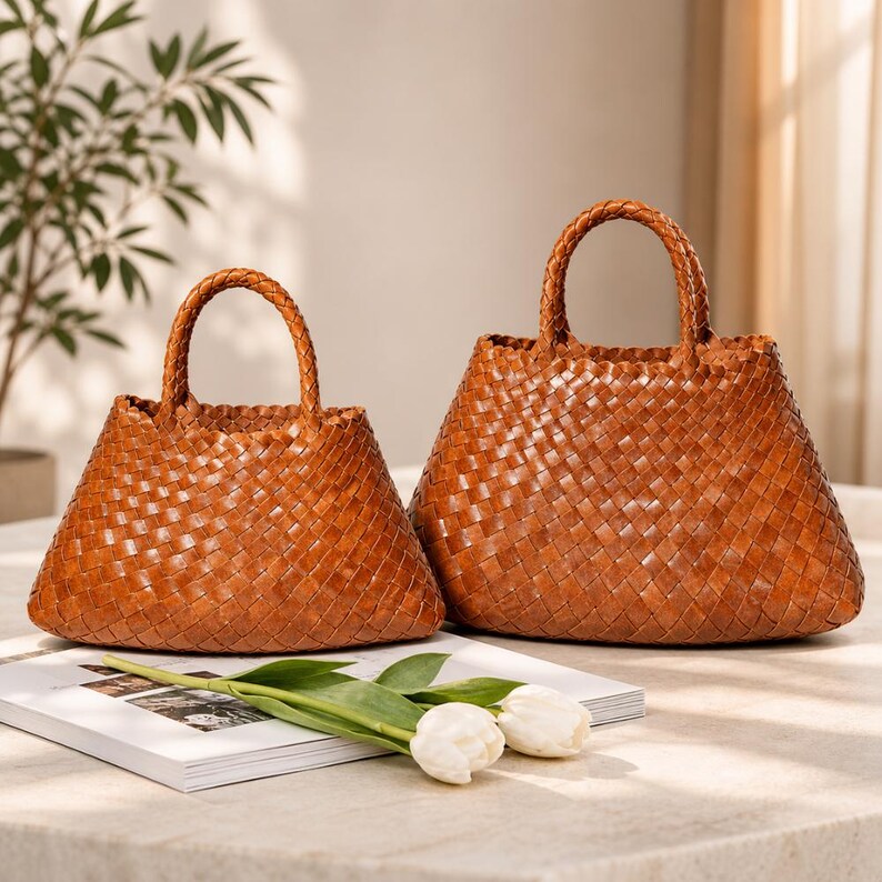 K&ouml;nnte beinhalten: Zwei braune, geflochtene Lederhandtaschen mit geflochtenen Griffen. Die Taschen haben ein korbartiges Webmuster. Eine Tasche ist kleiner als die andere. Wei&szlig;e Tulpen und eine Zeitschrift sind im Vordergrund.