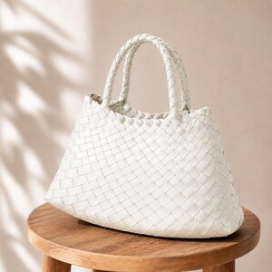 K&ouml;nnte beinhalten: Eine wei&szlig;e, gewebte Handtasche mit zwei geflochtenen Henkeln. Die Tasche hat eine trapezf&ouml;rmige Form und steht auf einem Holzschemel. Das Webmuster ist &uuml;ber die gesamte Oberfl&auml;che der Tasche sichtbar und erzeugt ein strukturiertes Aussehen.
