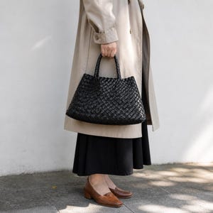 K&ouml;nnte beinhalten: Eine schwarze, gewebte Tragetasche mit strukturiertem Design. Die Tasche hat zwei Griffe und ein ger&auml;umiges Innenfach. Die Tasche wird von einer Person getragen, die einen beigen Trenchcoat und einen schwarzen Rock tr&auml;gt. Braune Lederloafers vervollst&auml;ndigen das Outfit.