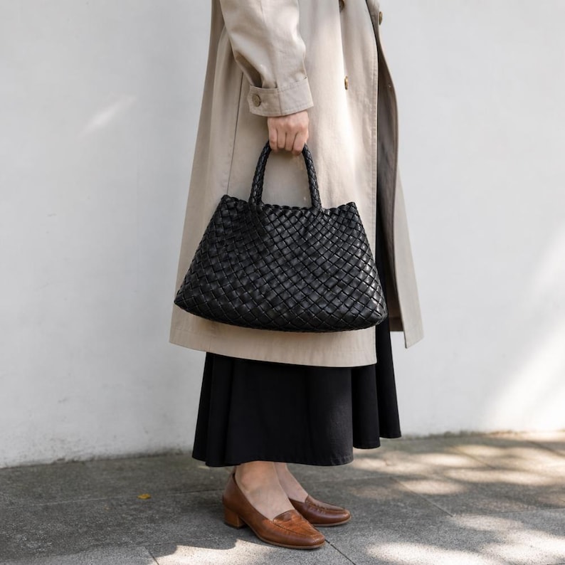 K&ouml;nnte beinhalten: Eine schwarze, gewebte Tragetasche mit strukturiertem Design. Die Tasche hat zwei Griffe und ein ger&auml;umiges Innenfach. Die Tasche wird von einer Person getragen, die einen beigen Trenchcoat und einen schwarzen Rock tr&auml;gt. Braune Lederloafers vervollst&auml;ndigen das Outfit.