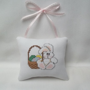 May include: White cross-stitch pillow featuring a cartoon Easter bunny holding a basket of colorful Easter eggs. The bunny is white with pink accents. A pink ribbon is attached to the top of the pillow for hanging. Easter-themed home decor.