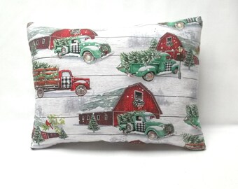 May include: Rectangular pillow with a winter scene featuring red barns, green trucks carrying Christmas trees, and a snowy landscape. The trucks have black and white checkered details. The background has a white wood grain pattern.