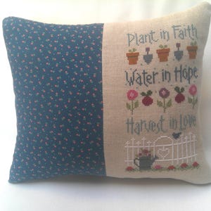 May include: A decorative pillow with a blue and white floral pattern on one side and a beige linen fabric with an embroidered quote on the other side. The quote reads "Plant in Faith, Water in Hope, Harvest in Love".