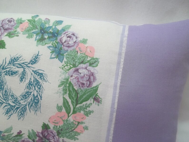 Puede incluir: Una almohada blanca con un dise&ntilde;o floral que presenta una corona de trigo y flores rosas, moradas y azules. La almohada tiene un respaldo de color lavanda.