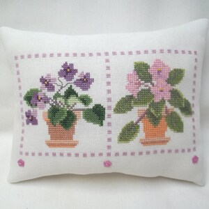 May include: A white pillow with two embroidered images of potted violet plants. One plant has purple flowers and the other has pink flowers.