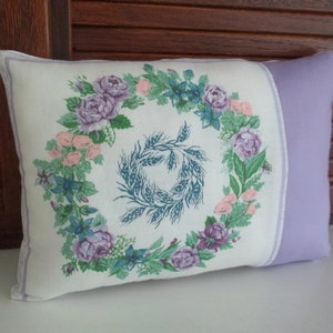 Puede incluir: Una almohada blanca con ribete lavanda presenta un dise&ntilde;o de corona floral con rosas moradas y rosas, hojas verdes y espigas de trigo azules.