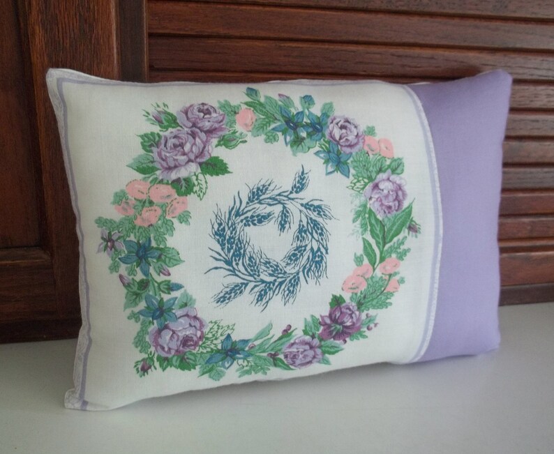 Puede incluir: Una almohada blanca con ribete lavanda presenta un dise&ntilde;o de corona floral con rosas moradas y rosas, hojas verdes y espigas de trigo azules.