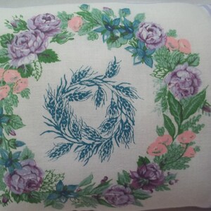 Puede incluir: Una funda de almohada blanca con un dise&ntilde;o floral morado y verde. El dise&ntilde;o floral presenta una corona de rosas, hojas y espigas de trigo. La funda de almohada tiene un borde morado claro.