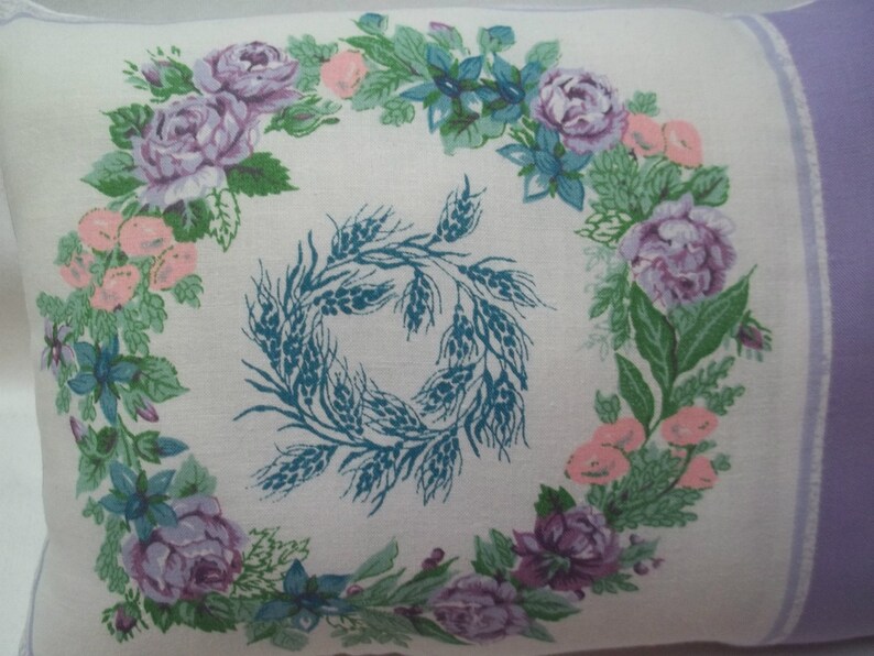 Puede incluir: Una funda de almohada blanca con un dise&ntilde;o floral morado y verde. El dise&ntilde;o floral presenta una corona de rosas, hojas y espigas de trigo. La funda de almohada tiene un borde morado claro.