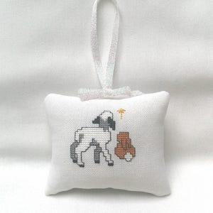 May include: White cross-stitch pillow ornament featuring a lamb and a brown bear. The lamb is gray and white, and the bear is brown. A gold star is above the bear. The ornament has a white ribbon for hanging.