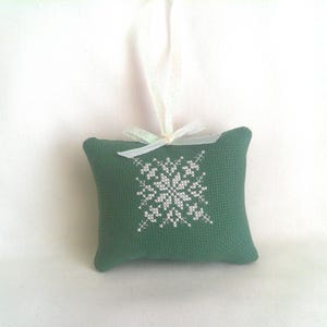 May include: A small, green, rectangular ornament with a white snowflake design embroidered on the front. A white ribbon is attached for hanging. The ornament is likely handmade and suitable for holiday decor.