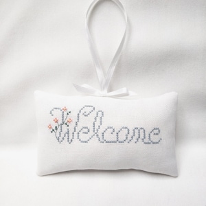 May include: A white fabric pillow with a gray embroidered "Welcome" message and a small pink floral design. The pillow has a white ribbon loop for hanging.