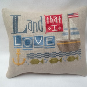 May include: A beige pillow with a cross-stitch design featuring the words "Land that I love" in blue and red, a sailboat with an American flag, an anchor, and fish.
