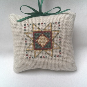 May include: A square, beige, woven fabric ornament featuring a cross-stitched star pattern in shades of yellow, dark green, and red. The ornament is topped with a dark green satin ribbon tied into a bow at the center for hanging purposes.