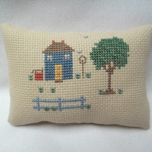 Pode incluir: Uma pequena almofada bege com um desenho de ponto cruz de uma casa azul com uma porta amarela, um balde vermelho, uma &aacute;rvore verde e uma cerca azul.