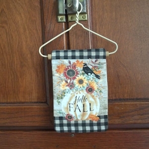 May include: A black and white checkered fabric flag with a white pumpkin and sunflowers. The pumpkin has the text "Hello Fall" on it. A black bird is perched on a branch above the pumpkin.