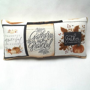 May include: A rectangular pillow with a white background and brown, black, and orange accents. The pillow features a patchwork design with three panels. The left panel has the text "Thankful grateful blessed" with a pumpkin design. The center panel has the text "Gather and be grateful" in black script. The right panel has the text "Gather together" with a pumpkin and fall leaves design.