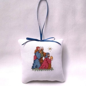 May include: A white, square, cross-stitched Christmas ornament featuring a Nativity scene with three figures and a star. The ornament has a blue ribbon for hanging. The figures are in shades of blue, red, and brown.