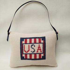 May include: A small, cream-colored pillow with a red, white, and blue embroidered design. The design features a square with stripes and the word "USA" in blue lettering. The pillow has a black ribbon loop for hanging.